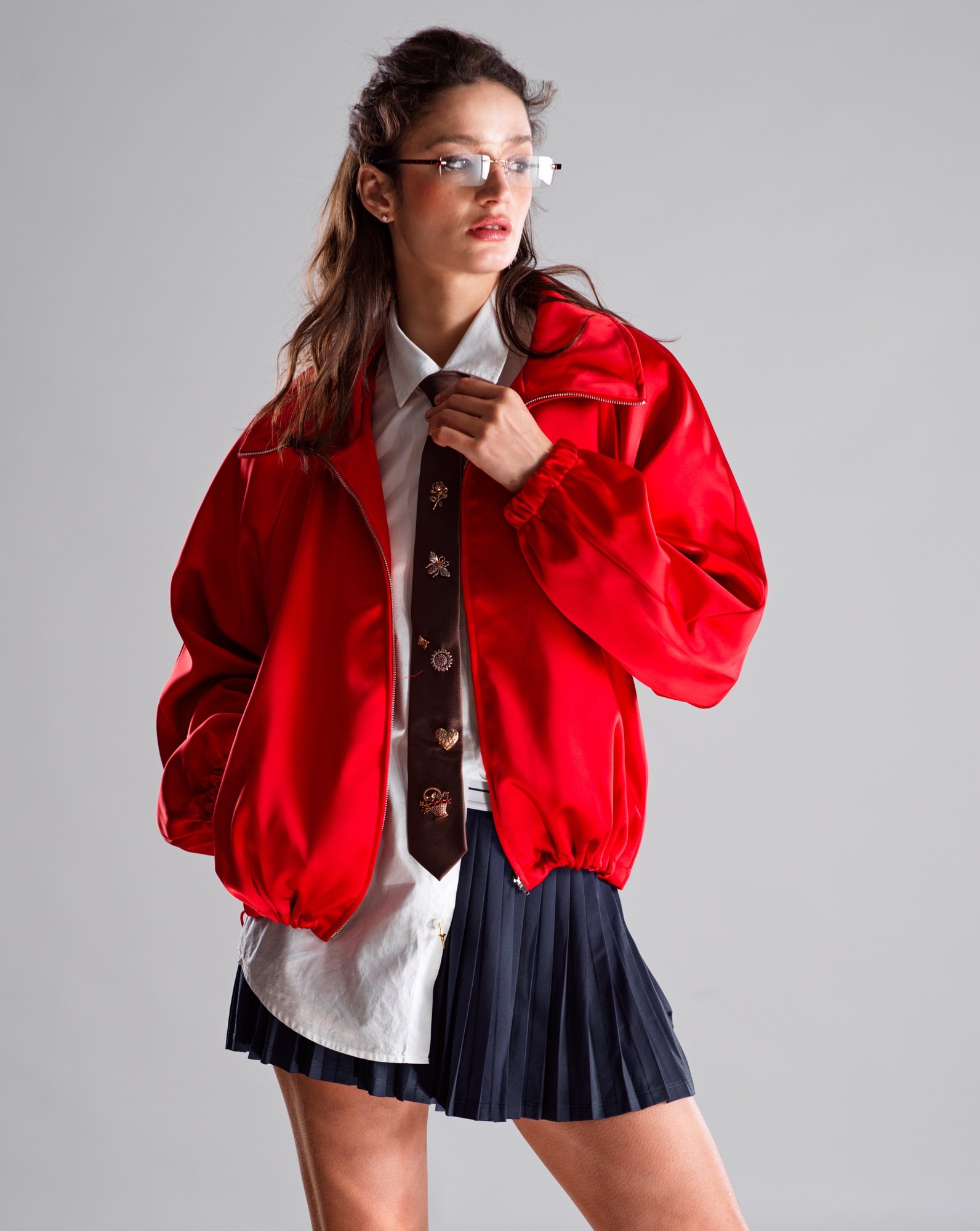 Model in an oversized red satin bomber jacket over a white shirt with a brown tie covered in gold pin badges, and a navy pleated skirt